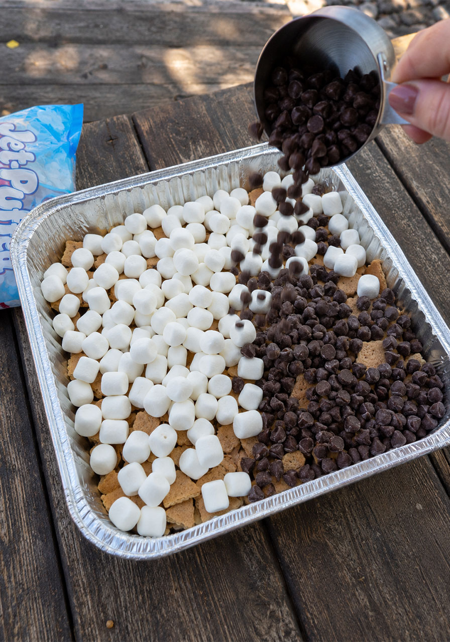 Adding mini marshmallows and chocolate chips to s'mores nachos in a foil pan