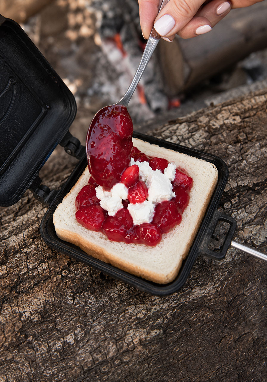 Spooning strawberry pie filling and cream cheese onto white bread inside a campfire pie iron