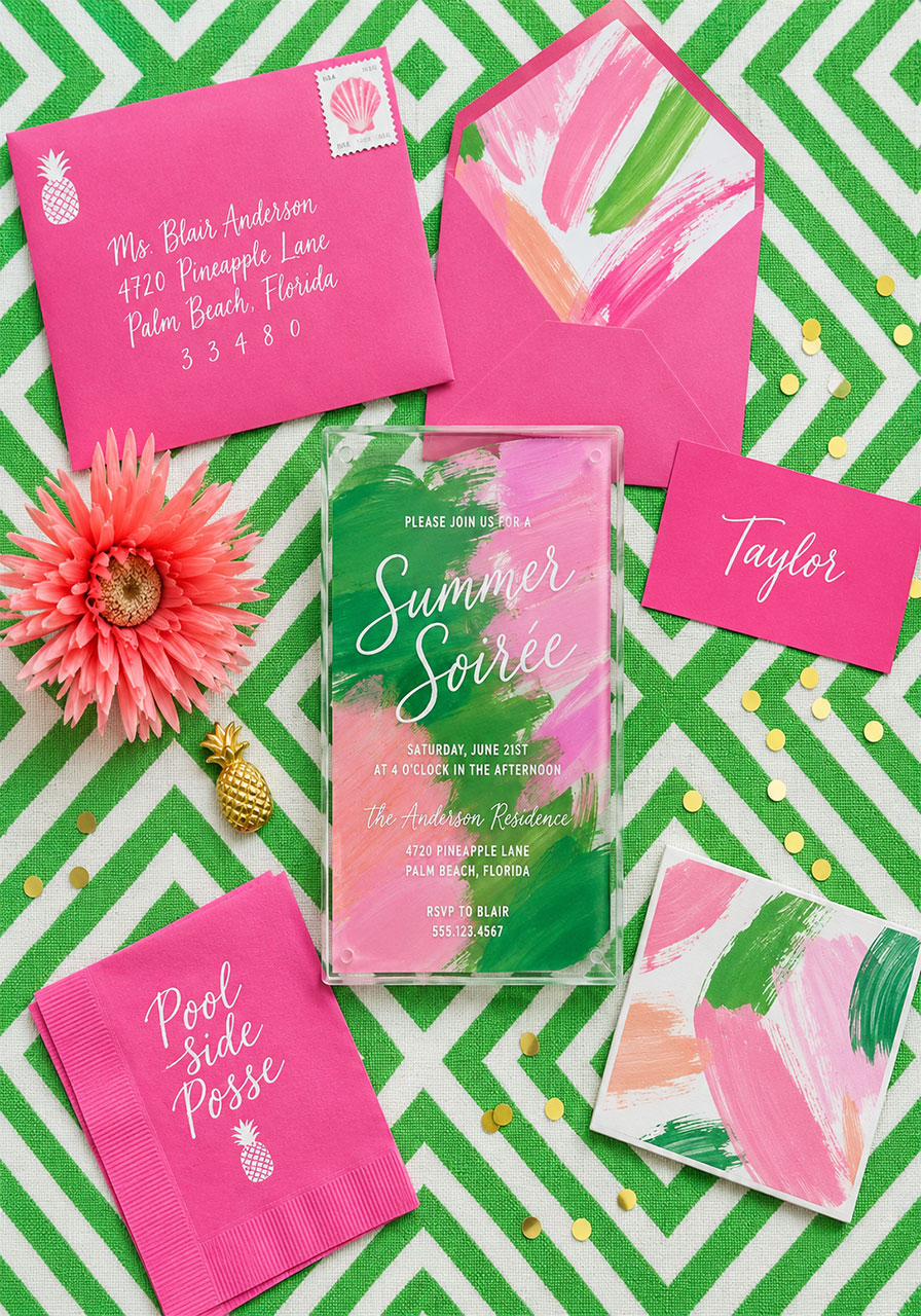 Flat-lay of pastel summer soirée acrylic invitation suite with pink envelopes, napkins, and gold pineapple charm