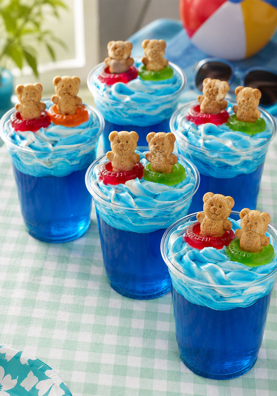 Swimming Bears Jello Cups pool party dessert – six blue Jello cups with whipped topping waves and Teddy Graham bear floaties on gingham tablecloth