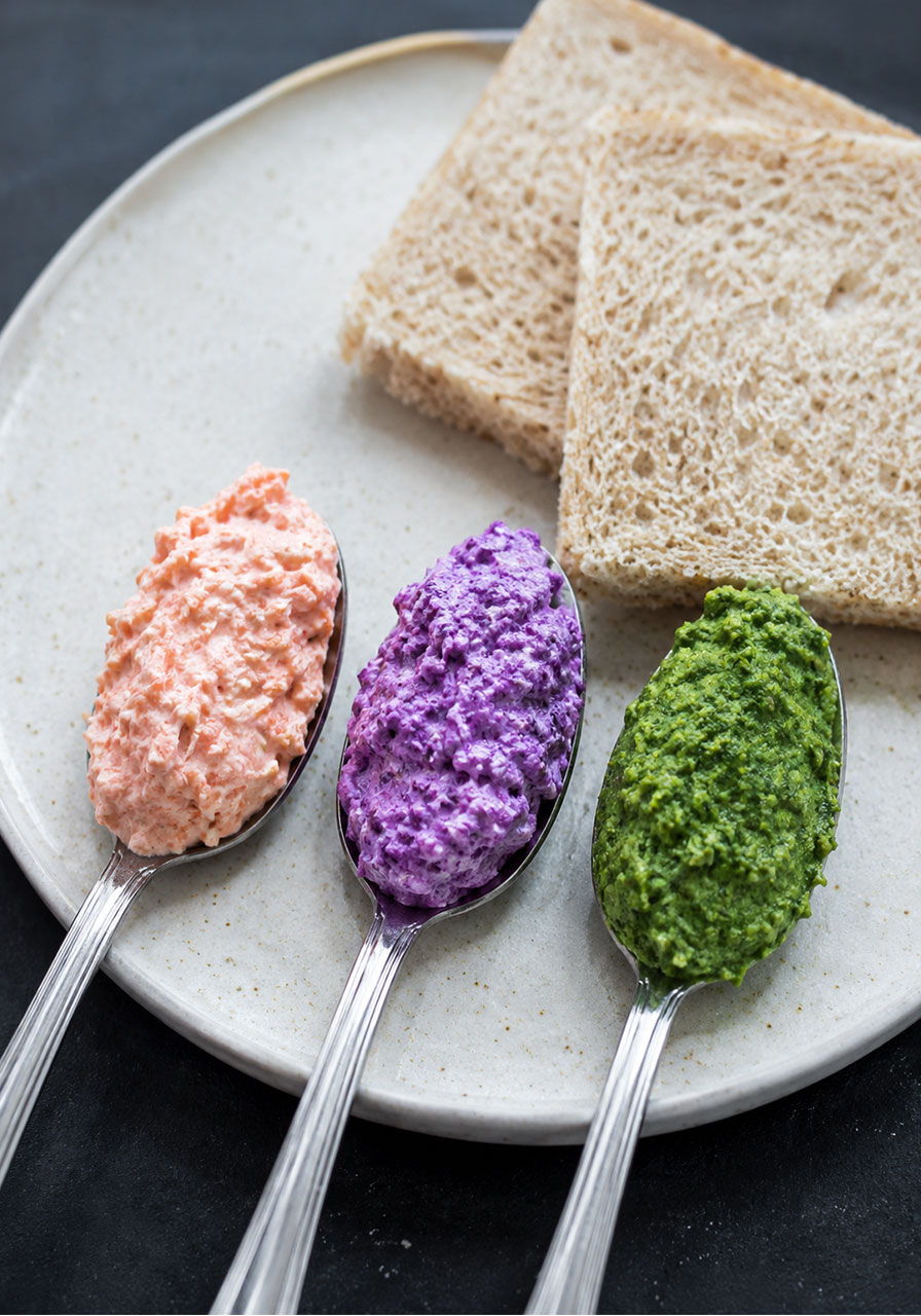 Three colorful sandwich spreads — orange carrot yogurt, purple cabbage yogurt, and green avocado cilantro dressing — on spoons next to bread slices