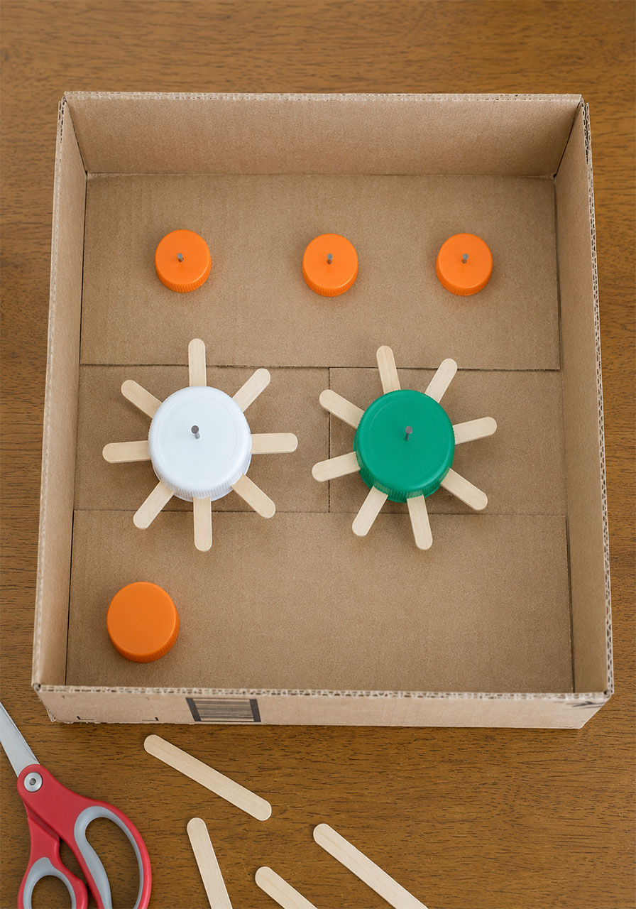 Small bottle caps glued as axle posts inside a cardboard box to let popsicle stick gears spin freely