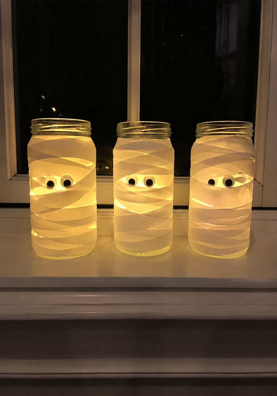 Three glowing mummy jar lanterns wrapped in white tape lit up at night on windowsill