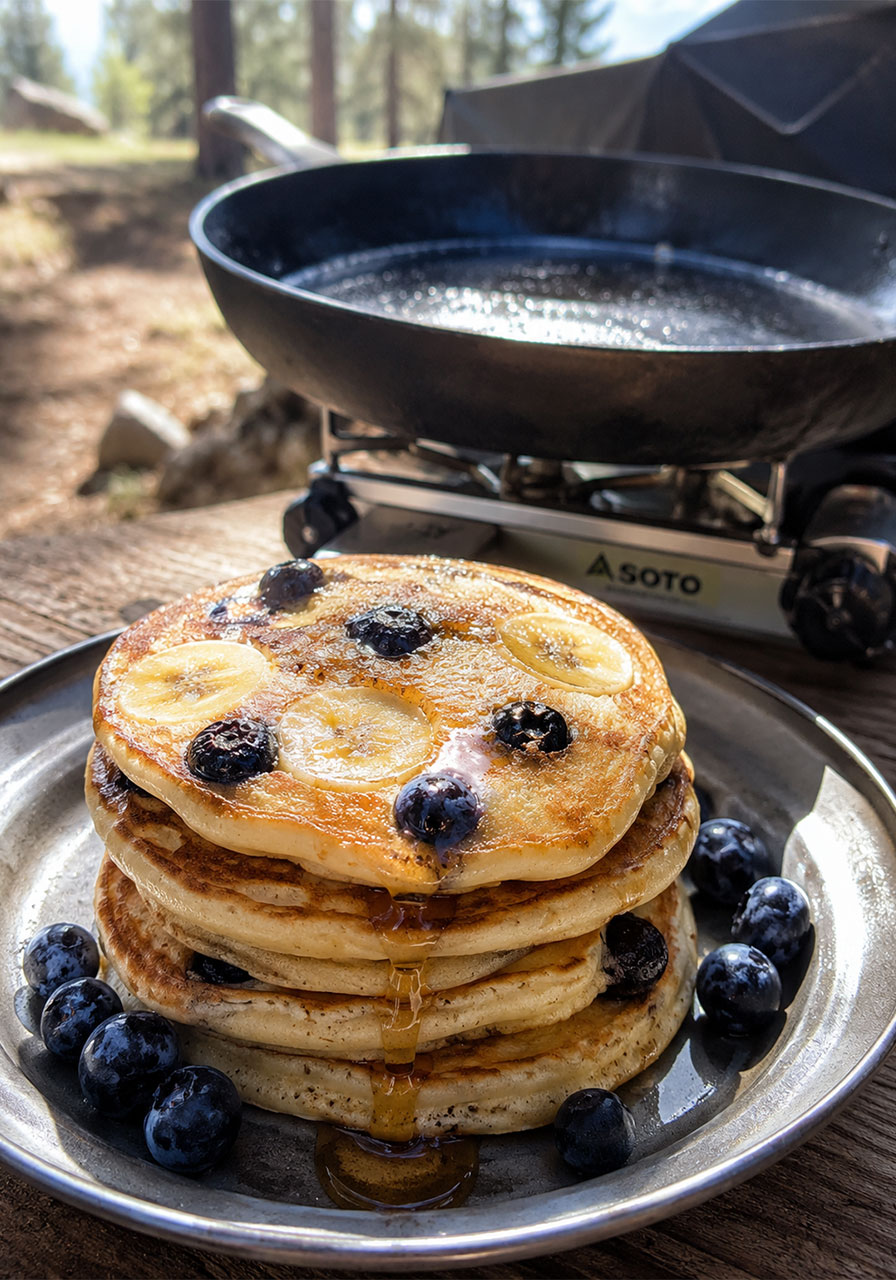 One pot camping breakfast: fluffy blueberry banana pancakes stacked on a camp plate with maple syrup
