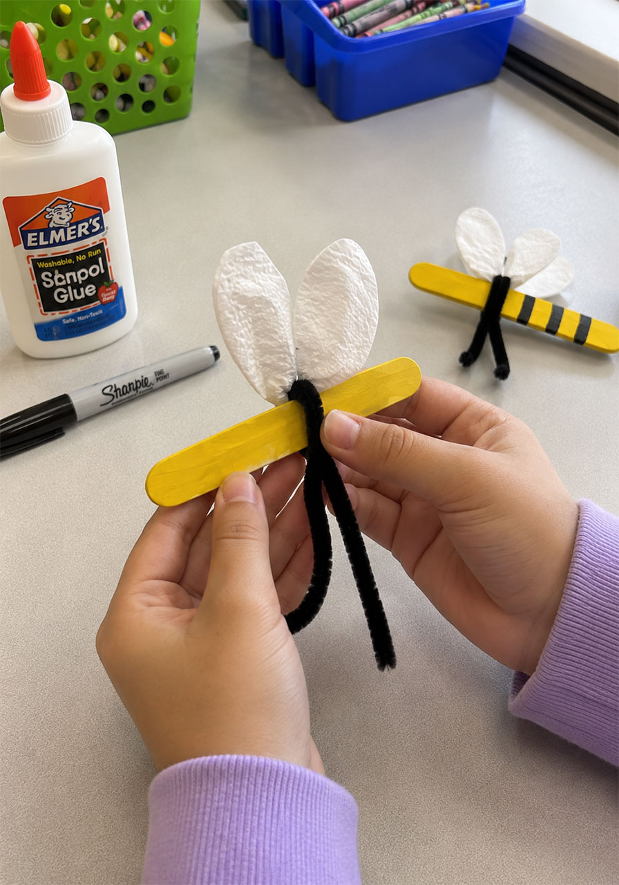 Child's hands wrapping a pipe cleaner around a craft stick to build a bee model for a pollination STEM activity