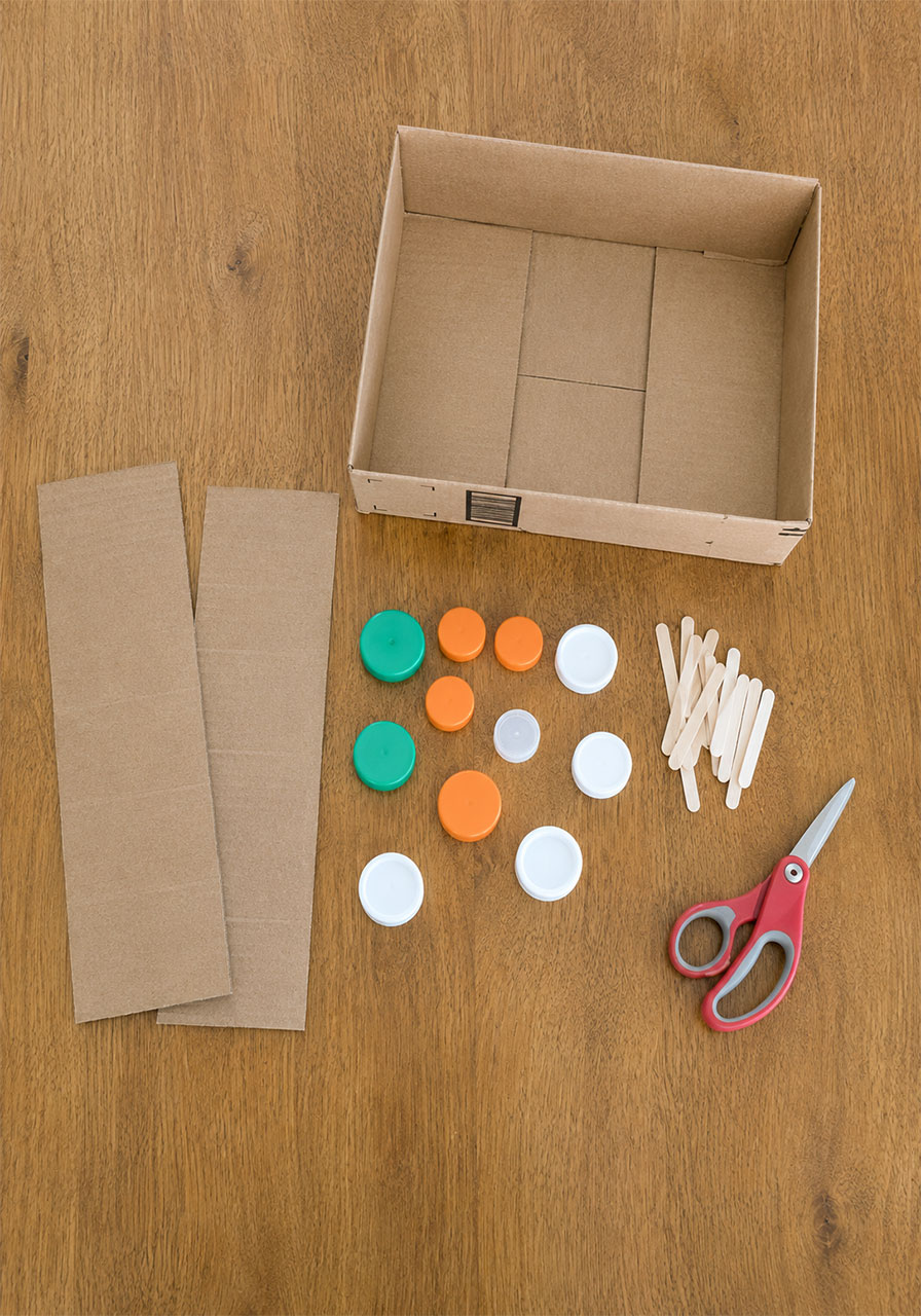 Materials needed for popsicle stick gears STEM activity including bottle caps, cardboard box, and craft sticks