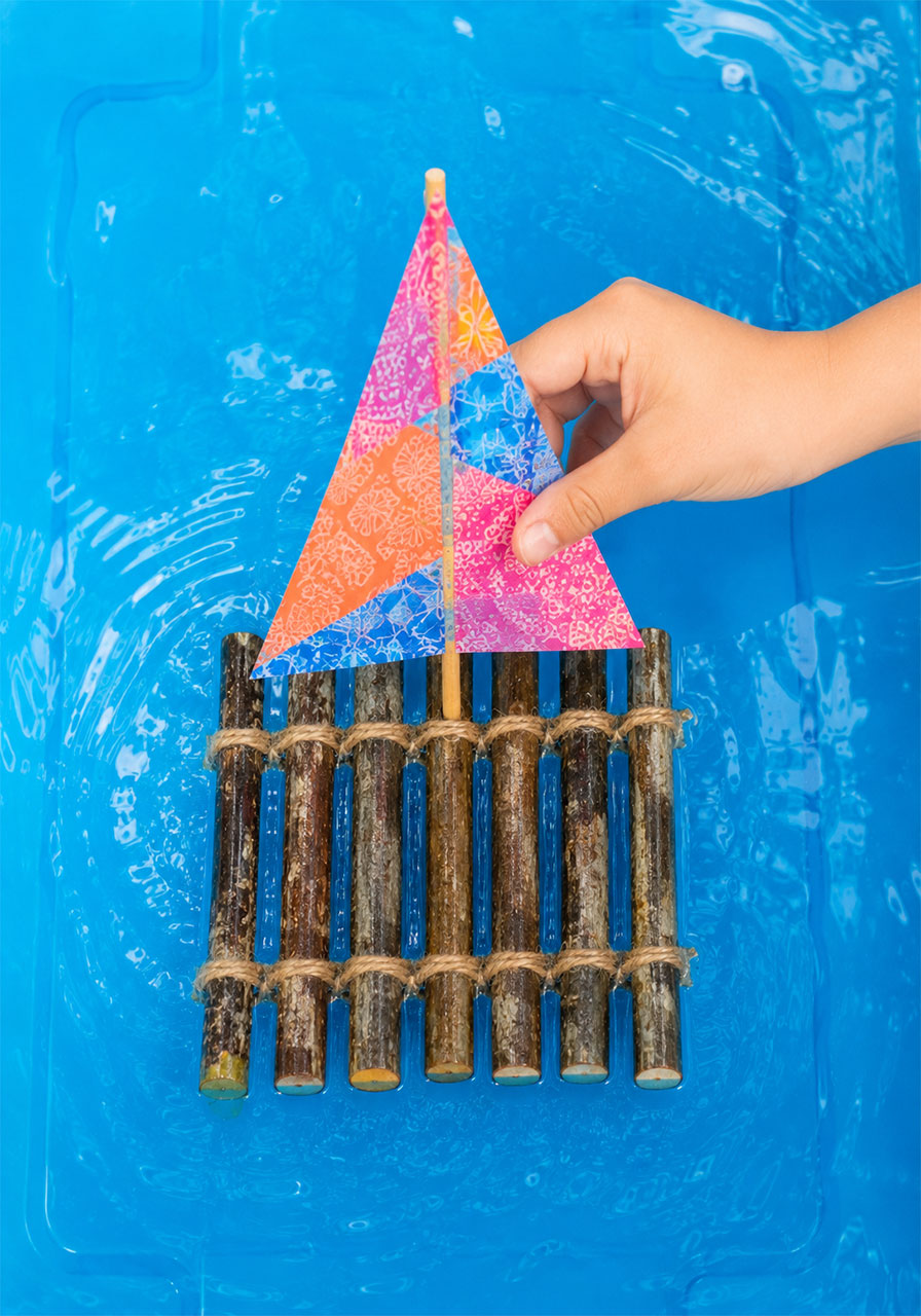Finished stick raft with colorful paper sail floating on blue water, a fun STEM challenge project
